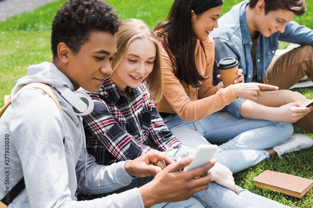 smiling and happy teenagers sitting on grass and looking at smartphone