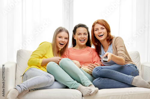 entertainment, leisure and friendship concept - three smiling teenage girls or friends with popcorn and remote watching tv at home