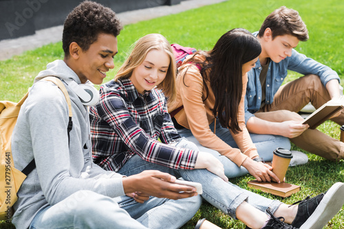 smiling and happy teenagers sitting on grass, looking at smartphone and reading book