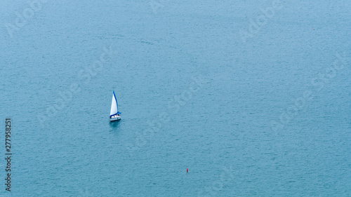 Small white and blue sailboat on calm, open blue water with small red buoy nearby