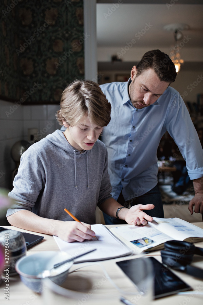 Father looking at son doing homework on table Father looking at son doing homework on table