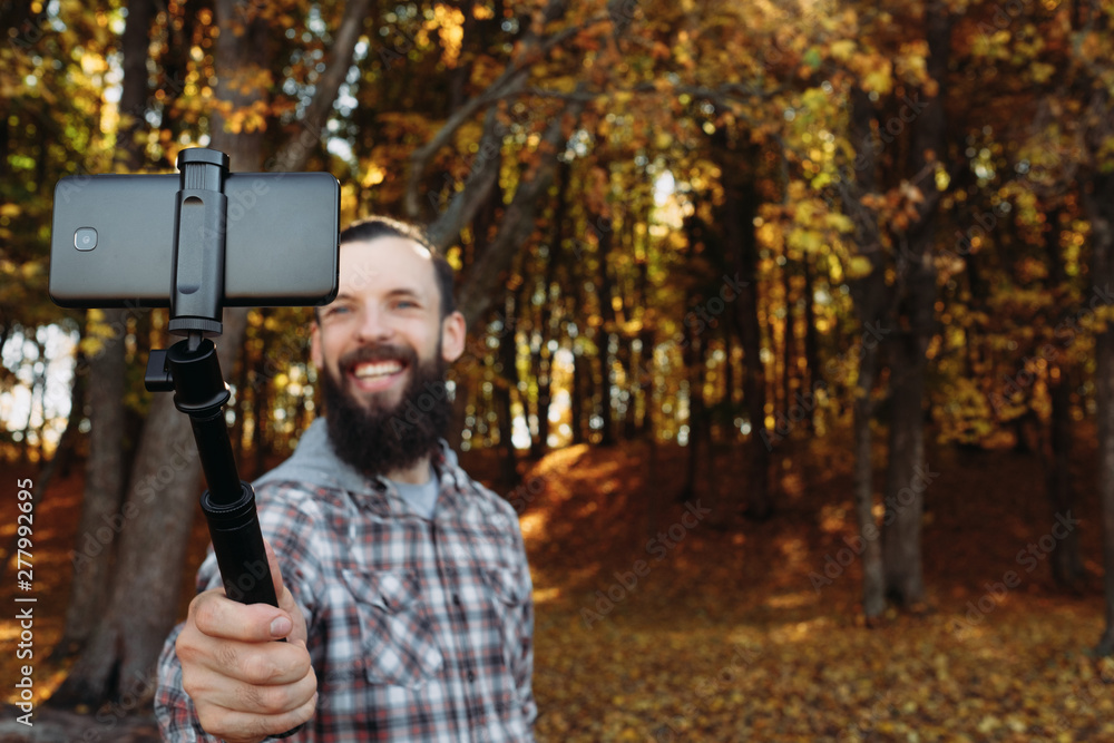 Fall nature leisure. Man using smartphone on stick to take selfie in autumn forest, smiling. Blur trees background. Copy space.