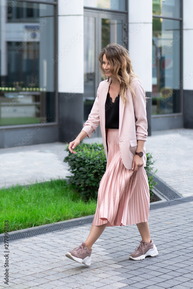 a girl in a peach jacket, a plush skirt and sneakers is walking along a stone street with a great mood. On the territory of a modern residential complex.