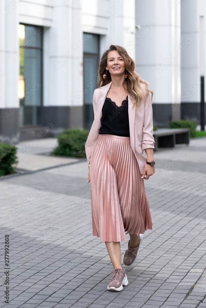 a young girl walks along the sidewalk in wide strides. Skirt plise, long jacket, sneakers. Long wavy hair. Very beautiful European in the summer walks down the street in a great mood. a young girl walks along the sidewalk in wide strides. Skirt plise, long jacket, sneakers. Long wavy hair. Very beautiful European in the summer walks down the street in a great mood.