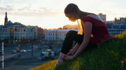 Young ballerina fastens the tug on her pointe sitting on a green hill in the middle of the city. photo