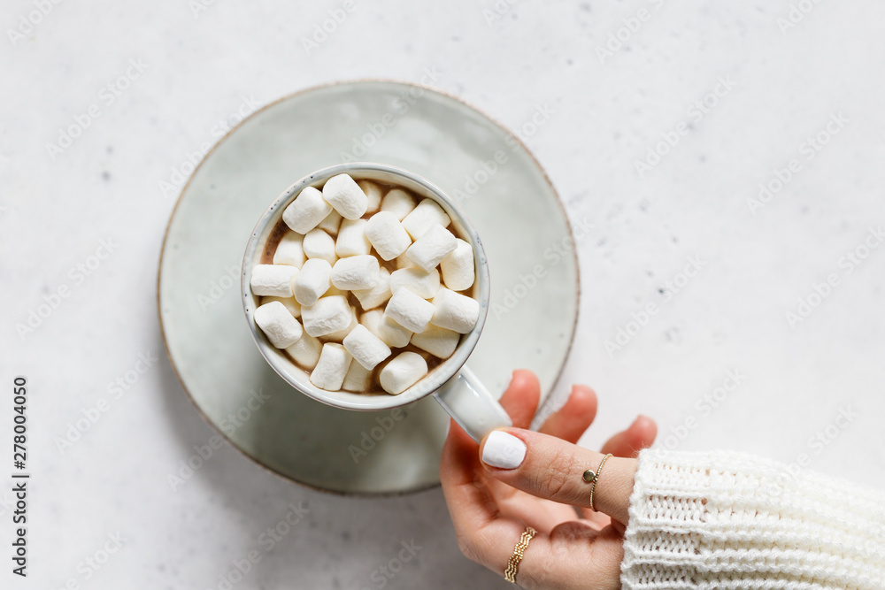 Girl's hand holds a cup of hot chocolate with marshmallow over textured white background. Minimalist style, top view, copy space. Girl's hand holds a cup of hot chocolate with marshmallow over textured white background. Minimalist style, top view, copy space.