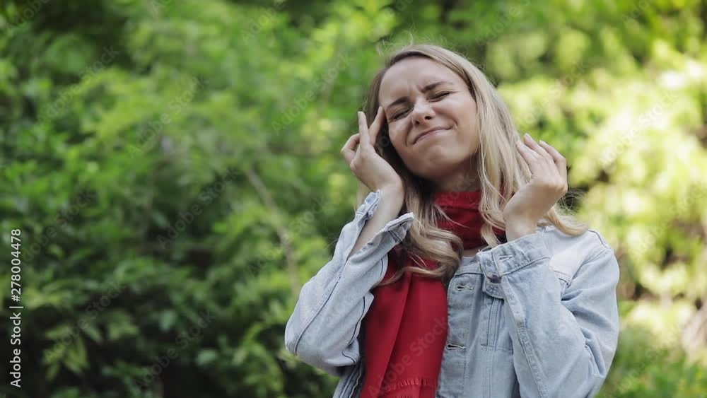 Young woman wearing red scarf with headache standing in the park. Girl feel ill unhappy and with migraine touching her temples. Dreadful migraine. Young woman wearing red scarf with headache standing in the park. Girl feel ill unhappy and with migraine touching her temples. Dreadful migraine.