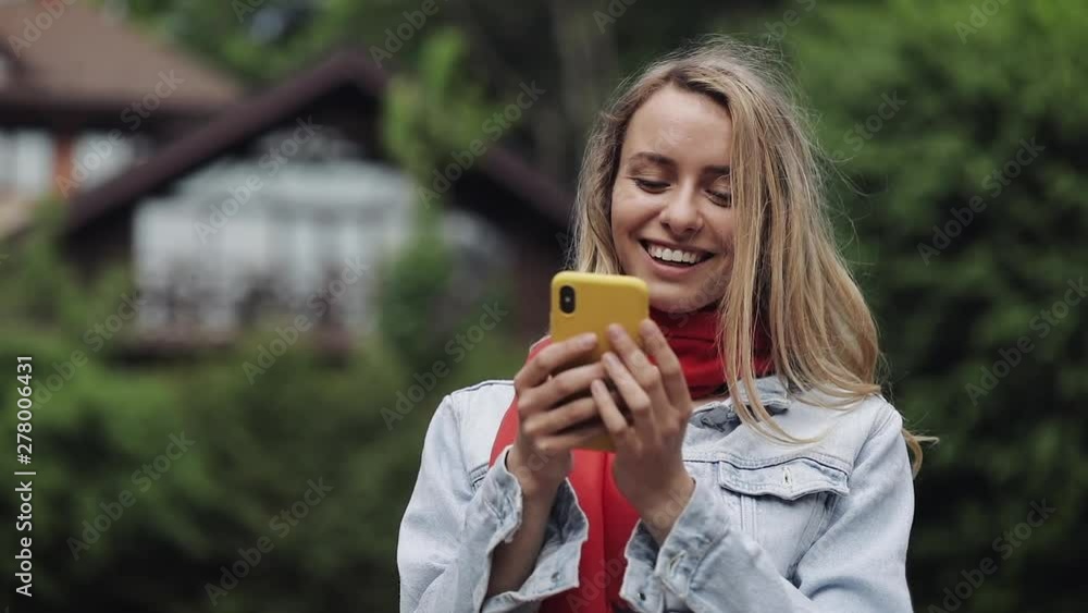 Portrait of young beautiful woman using app on smartphone, smiling and texting on mobile phone. Woman wearing in the red scarf standing near a country house. Portrait of young beautiful woman using app on smartphone, smiling and texting on mobile phone. Woman wearing in the red scarf standing near a country house.