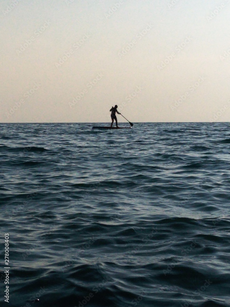 a small black silhouette of a swimmer on a sub Board with an oar on a background of light gray sky and Indigo water. small waves on the surface of the sea, glare, soft diffused light of the setting su