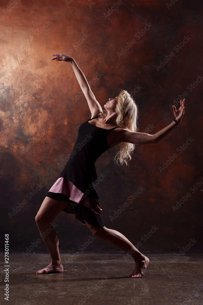 Photo of dancing blonde looking back in black dress on brown background in studio