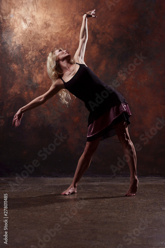 Full-length photo of dancing blonde girl looking up in short black dress on brown background
