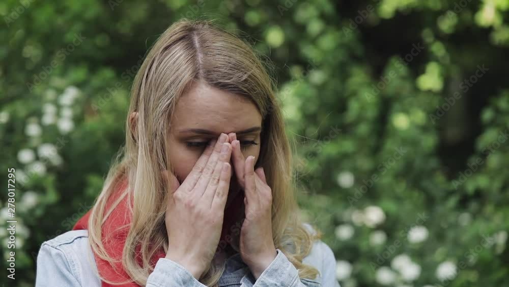 Young beautiful woman rubs her eyes standing against the background of flowering trees. Allergy, rhinitis, eye diseases, sinus pressure pain. Young beautiful woman rubs her eyes standing against the background of flowering trees. Allergy, rhinitis, eye diseases, sinus pressure pain.