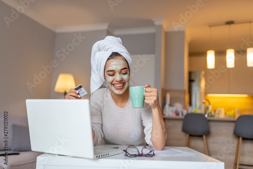 Woman with cosmetic face mask shopping online from her home. Leisure time at home. pPortrait of beautiful woman with facial taking a coffee at home while use internet with modern laptop computer photo