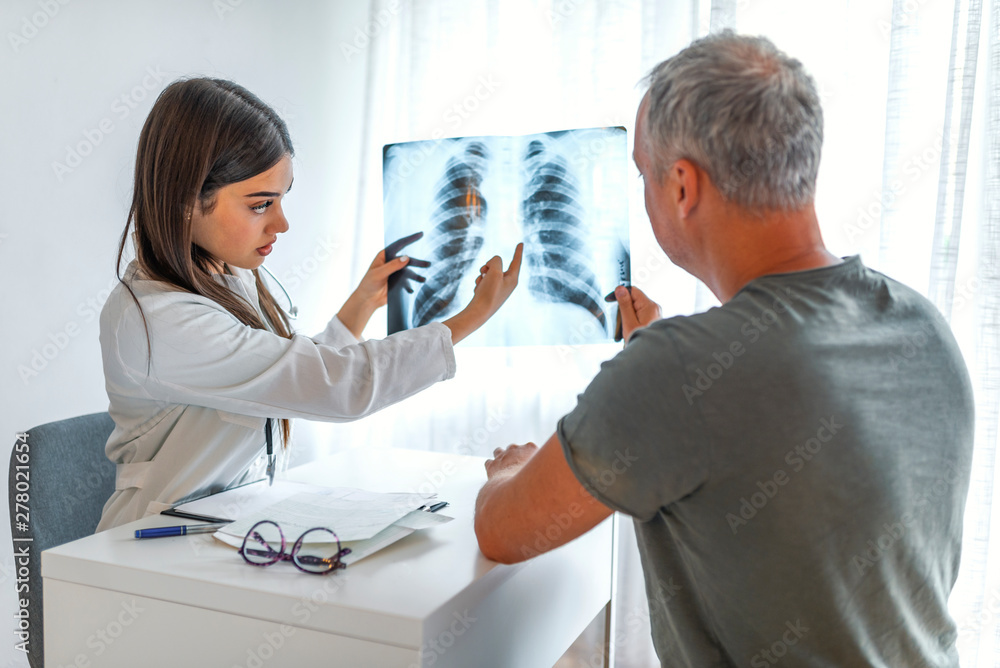 Young doctor talking to a patient in her cabin. Doctor showing x-ray to patient. Doctor and patient looking at Xray in medical office. Doctor shows the patient chest x-ray Young doctor talking to a patient in her cabin. Doctor showing x-ray to patient. Doctor and patient looking at Xray in medical office. Doctor shows the patient chest x-ray