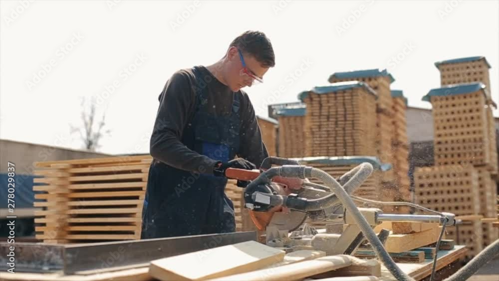 Close up of a young carpenter worker wearing construction goggles and uniform using wood cutting machine. man cuts wooden planks. Slow motion. Close up of a young carpenter worker wearing construction goggles and uniform using wood cutting machine. man cuts wooden planks. Slow motion.