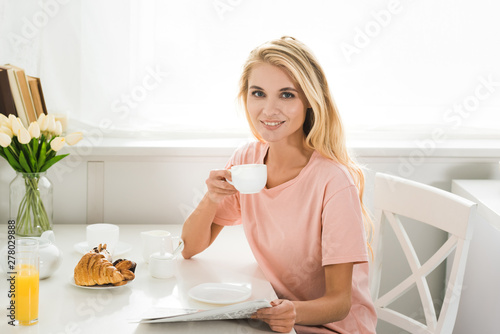 beautiful happy woman with coffee cup and newspaper at kitchen table in morning