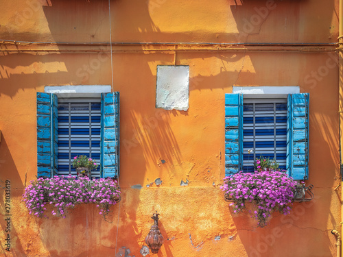 Two windows with flowerboxes and open blue shutters on a orange shabby plastered facade of the ancient building. Flowers in boxes on a wall of the old house with open windows. photo