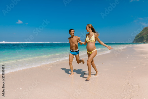 Happy running couple. Pregnant woman with husband expecting baby at tropical beach