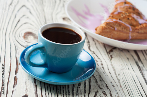 Full ceramic cup of hot black coffee near baked sweet bun in form of triangle with purple cream lies on white weathered wooden table