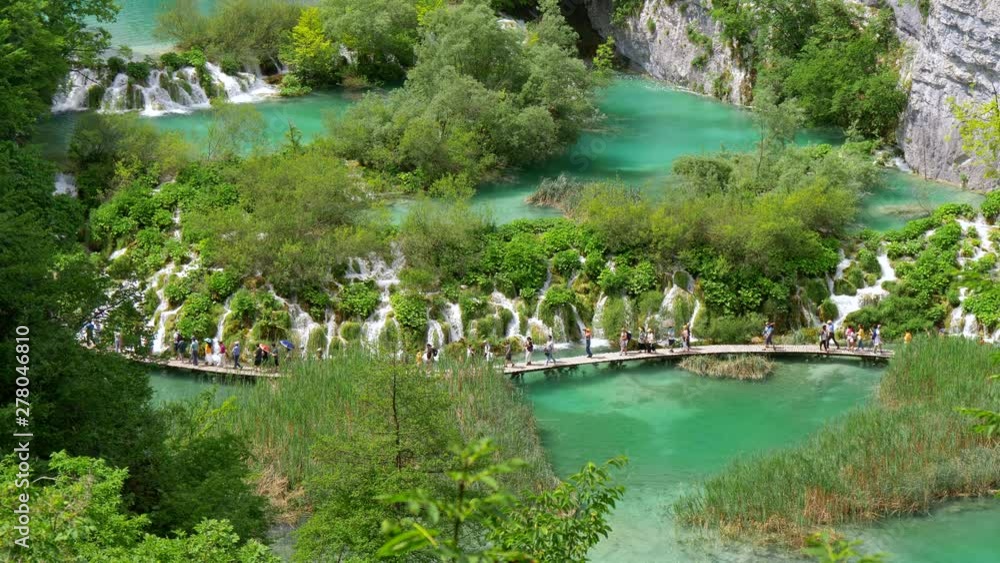 Tourists walking the boardwalk over a Plitvice lake in Plitvice Lakes National Park, Croatia. There are beautiful small cascades. Turquoise waters of the lake are surrounded with rich green flora. UHD Tourists walking the boardwalk over a Plitvice lake in Plitvice Lakes National Park, Croatia. There are beautiful small cascades. Turquoise waters of the lake are surrounded with rich green flora. UHD
