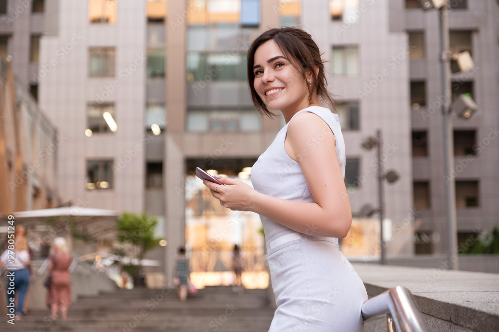 Beautiful brunette business woman in grey smark casual dress working on a mobile phone in her hands outdoors. European city on background. copy space