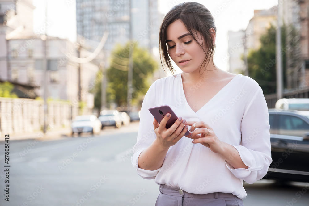 Fototapeta premium Beautiful brunette business woman in white skirt and grey suit trousers working on a mobile phone in her hands outdoors. European city on background. copy space