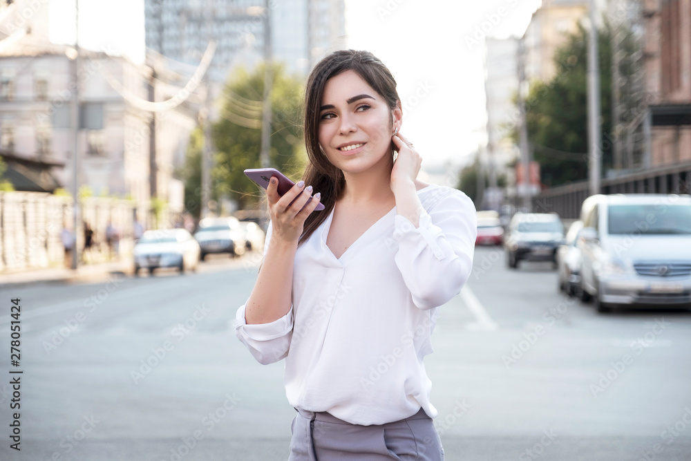 Beautiful brunette business woman in white skirt and grey suit trousers records voice messages on a mobile phone in her hands outdoors. European city on background. copy space Beautiful brunette business woman in white skirt and grey suit trousers records voice messages on a mobile phone in her hands outdoors. European city on background. copy space