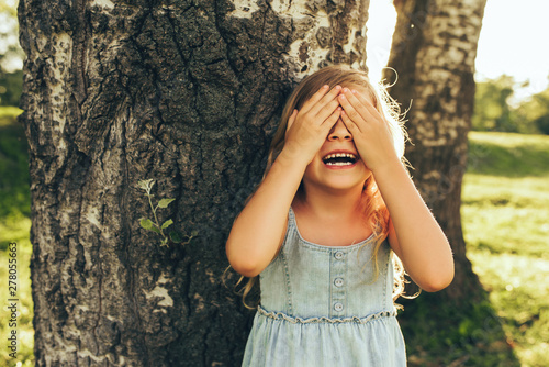 Foto Horizontal outdoor image of smiling little girl covering her eyes with both hands, playing hide and seek standing next a big tree