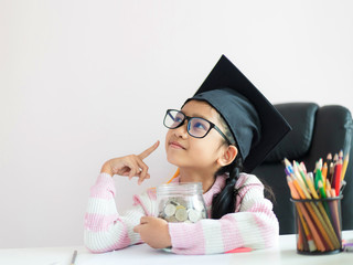 Little Asian girl wearing graduate hat hugging clear glass jar piggy bank and smile with happiness for money saving to wealthness succesful in the future of education concept