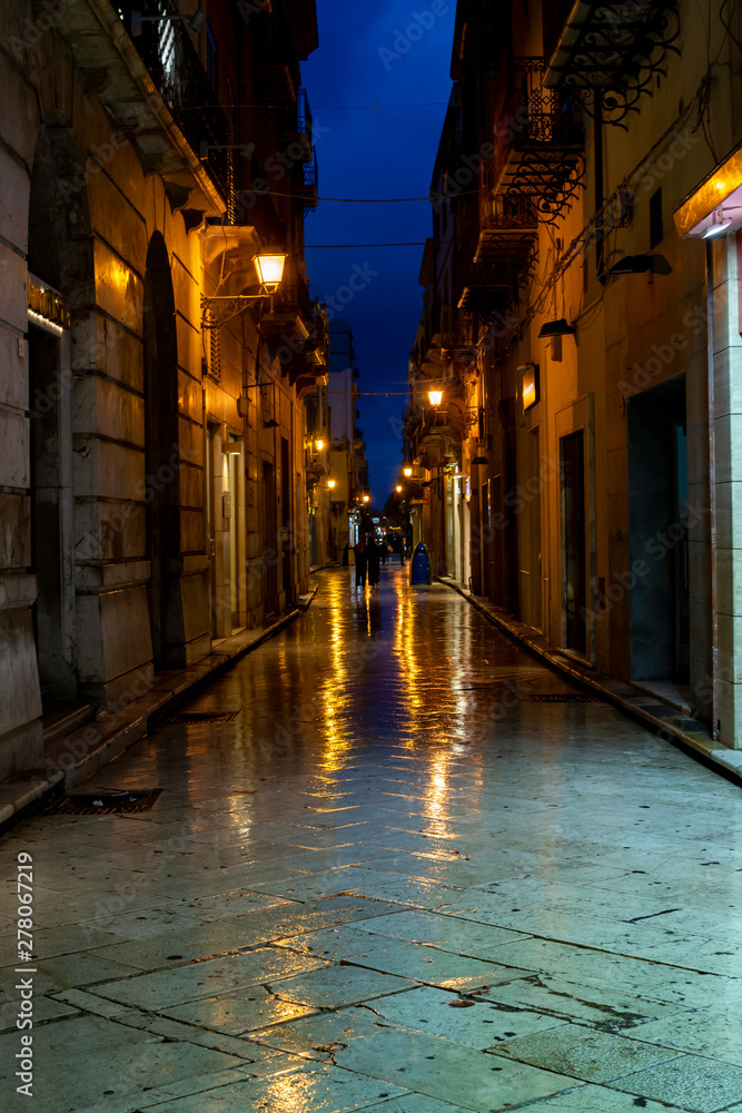 Old street in Marsala at night in rain with reflection of street lights on water, Sicily, Italy