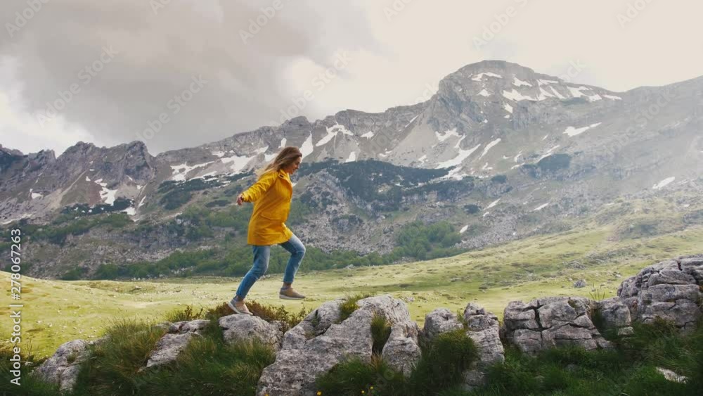 Young cheerful woman in a yellow raincoat running and having some fun on rocks with beautiful mountains background, slow motion