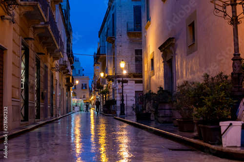 Fototapeta Naklejka Na Ścianę i Meble -  Old street in Marsala at night in rain with reflection of street lights on water, Sicily, Italy