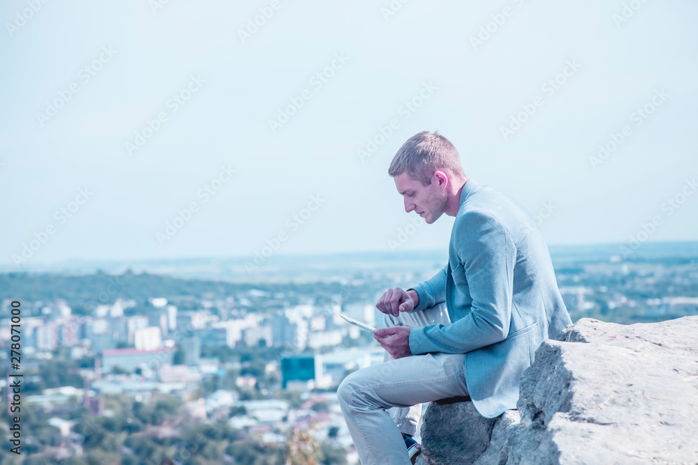 Concept: studying and working anytime and anywhere. Young handsome successful man is sitting on the stone and working with digital tablet over city from the mountains above.
