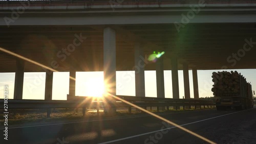 timber truck carries wood logs against the background of a modern bridge and a sunset, cargo transportation concept, trucking industry