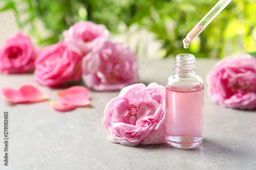 Dripping rose essential oil into bottle with pipette and fresh flowers on grey table, space for text
