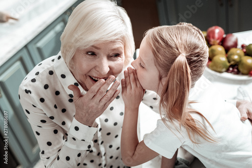 Emotional granny touching her mouth while little girl sharing secrets with her