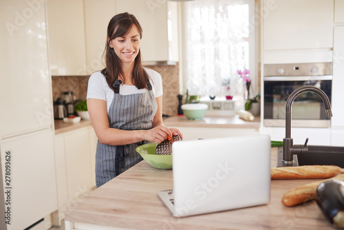 Young Caucasian brunette in apron grating carrots in bowl, preparing dinner and looking at lapotp while standing in kitchen