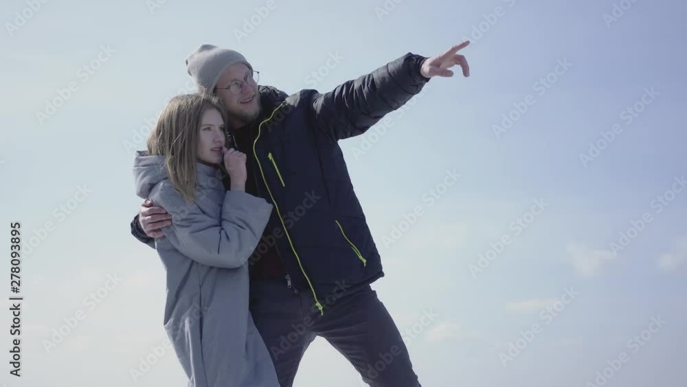 Beautiful blond couple pointing away hugging each other. The bearded man in glasses and attractive woman admiring nature in the winter. Tourists travel together Beautiful blond couple pointing away hugging each other. The bearded man in glasses and attractive woman admiring nature in the winter. Tourists travel together