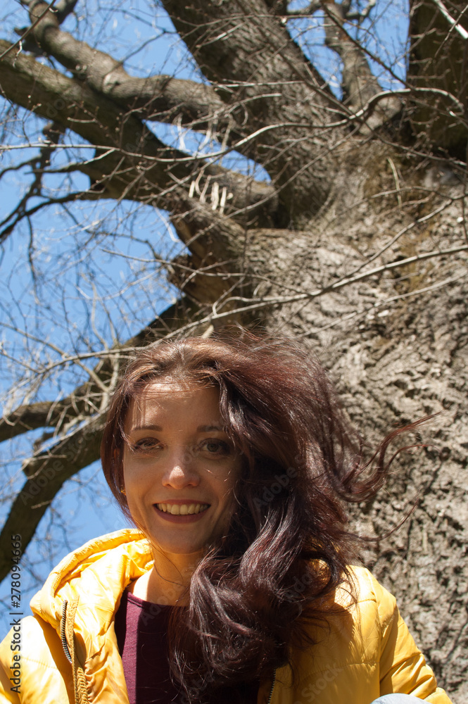 Close up outdoos portrait of young smilling girl in yellow jacket sitting near big old tree in the park during sunny day in early spring Close up outdoos portrait of young smilling girl in yellow jacket sitting near big old tree in the park during sunny day in early spring