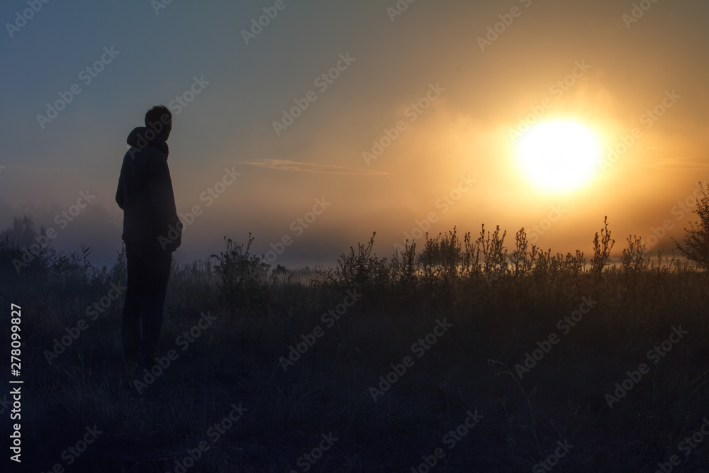 Silhouette of the traveler against fog landscape over a flower meadow, the first rays of dawn and dark silhouettes of trees against a sunrise, selective focus