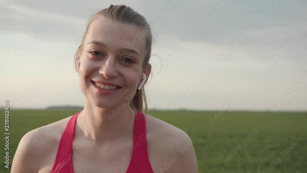 Young fitness sport woman with long hair use headphone look at camera smile happy healthy marathon workout beautiful nature road sport woman jogging park portrait slow motion Young fitness sport woman with long hair use headphone look at camera smile happy healthy marathon workout beautiful nature road sport woman jogging park portrait slow motion