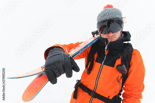 Portrait of a professional athlete skier in an orange jacket wearing a black mask and with skis on his shoulder looks into the camera. Gray background during snowstorm © yanik88