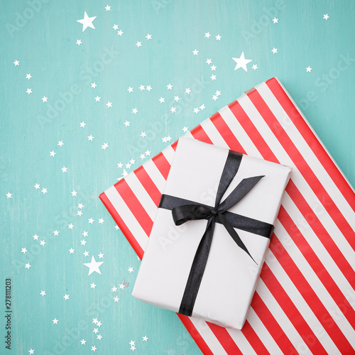 Closeup on gifts wrapped in white and striped red paper with black ribbon. Gifts lie on a turquoise wooden background covered with silver stars. Stylish holiday and christmas concept