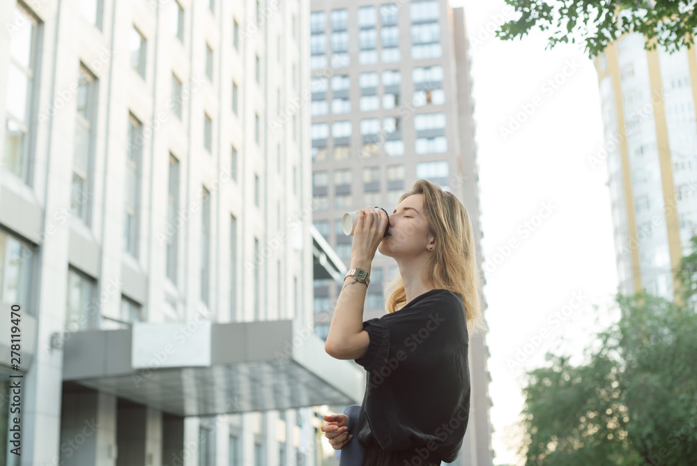 Cheerful stylish businesswoman drinking coffee outside on urban background. Attractive young woman enjoying coffee outdoors on the street near the office building and skyscraper. Cheerful stylish businesswoman drinking coffee outside on urban background. Attractive young woman enjoying coffee outdoors on the street near the office building and skyscraper.