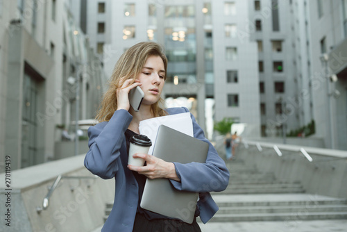 Obraz na plátne Young confident business woman walking in hurry downstairs, holding documents, laptop, coffee cup, talking on mobile phone on city street in front of modern office building