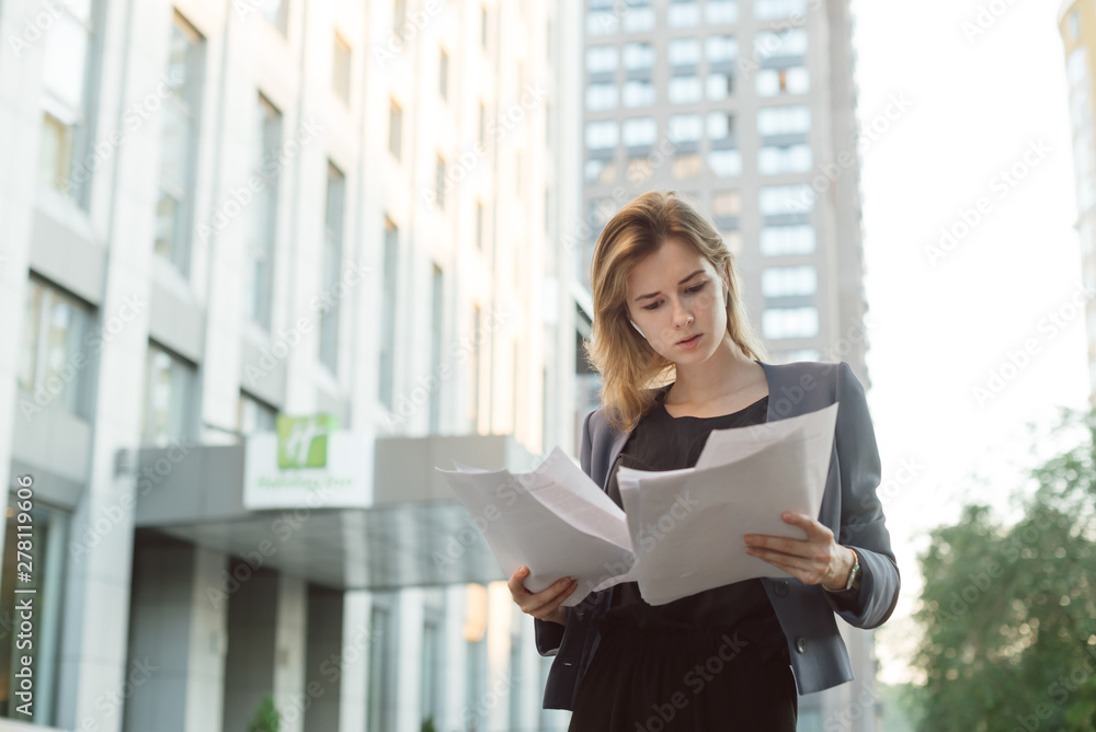 Concentrated caucasian financial adviser viewing business papers outdoors. Busy young businesswoman examining data in documents. Report concept. Office worker reading the documents on the street.