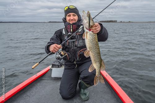 Pike fishing. Happy fisherman with big fish trophy at the boat with tackles