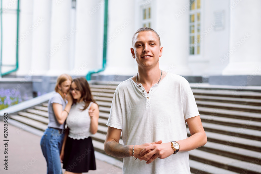 Handsome student standing in front of college building while girls are gossiping about him, flirts and staring at him Handsome student standing in front of college building while girls are gossiping about him, flirts and staring at him