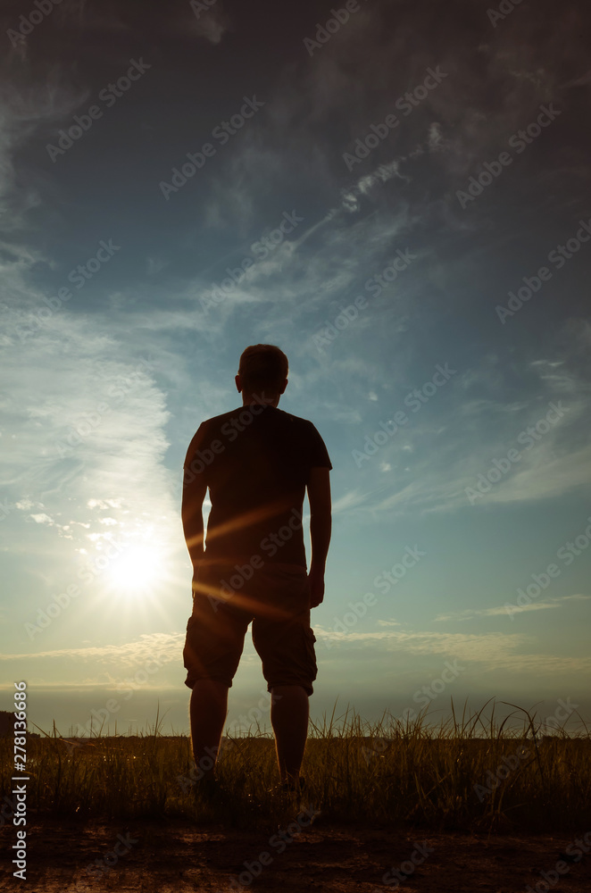 Silhouette of a young man against the backdrop of the setting sun and sky. The concept of loneliness, freedom, choice. Man of dense build in a t-shirt and shorts. Place for text, minimalism.