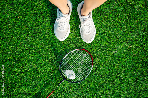 Red badminton rackets and shuttlecock lie on the artificial grass of a sports stadium, white sneakers stand next to them. Amateur badminton set. Concept of summer hobby, outdoor sports, entertainment.